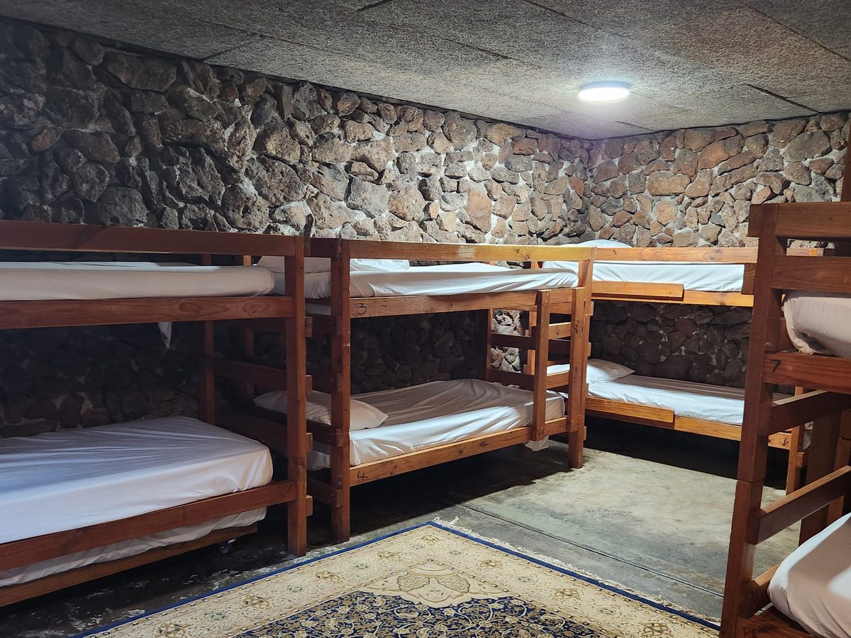A homestead bunk room with rows of timber bunks made up with white linen against a stone wall